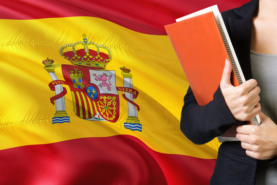 Learning Spanish Language Concept. Young Woman Standing With The Spain Flag In The Background. Teacher Holding Books, Orange Blank Book Cover.