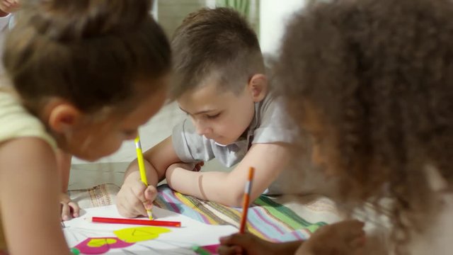 Close up shot of little multiethnic boys and girls lying on the floor and drawing with colored pencils on handmade collage