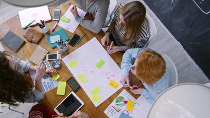 Zoom out top view of team of young colleagues sitting together at office table and drawing business scheme on paper while working together on creative project - Powered by Adobe