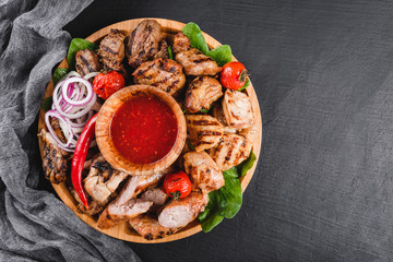 Assorted delicious grilled meat and sausages with tomatoes and bbq sauce on cutting board over black stone background.