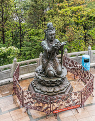 Hong Kong, China - March 7, 2019: Lantau Island. One of the Six Devas offers fruit to Tian Tan Buddha. Bronze statue seen from higher up with green foliage in back.