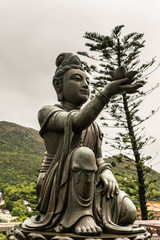 Hong Kong, China - March 7, 2019: Lantau Island. Closeup, One of the Six Devas offers flower to Tian Tan Buddha. Bronze statue seen from front with green foliage and rainy sky in back.