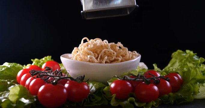 On a black background, vegetables and noodles on a wooden table, the whole dish is rubbed on top with parmesan cheese and mixed noodles. Concept of: Grater, Cheese, Noodles, Cherie Tomatoes, Salad.
