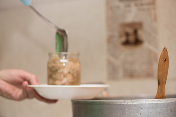 Filling a glass jar with boiled meat. Cooking pork stew at home, process close-up. Wooden spoon sticks out of pan