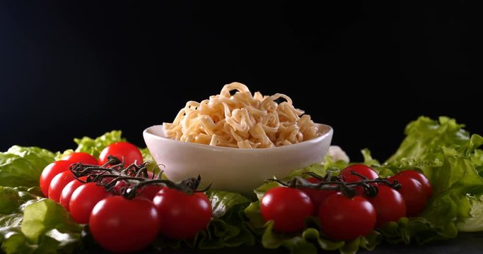 On a black background, vegetables and noodles on a wooden table, the whole dish is rubbed on top with parmesan cheese and mixed noodles. Concept of: Grater, Cheese, Noodles, Cherie Tomatoes, Salad.