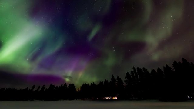 Time-lapse of fast moving vivid green and purple Aurora (Northern Lights) above a cozy cabin and reflecting on a frozen Canadian lake with spikes of blue and stars