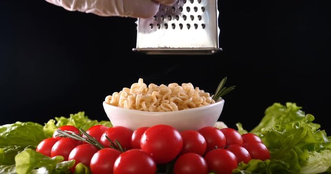 On a black background, vegetables and noodles on a wooden table, the whole dish is rubbed on top with parmesan cheese and mixed noodles. Concept of: Grater, Cheese, Noodles, Cherie Tomatoes, Salad.