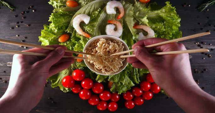 Background of lettuce leaves and a wooden table, a ready-made dish of vegetable noodles and a sea of products are being detained and then tried (eaten) with Chinese sticks. Concept: Shrimps, Vegetable
