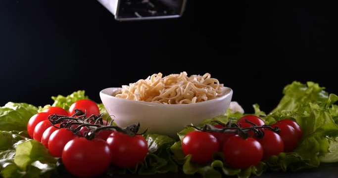 On a black background, vegetables and noodles on a wooden table, the whole dish is rubbed on top with parmesan cheese and mixed noodles. Concept of: Grater, Cheese, Noodles, Cherie Tomatoes, Salad.