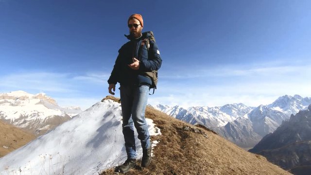 Happy traveler bearded man photographer in sunglasses with a camera and a backpack goes uphill on a background of snow-capped peaks on a sunny day