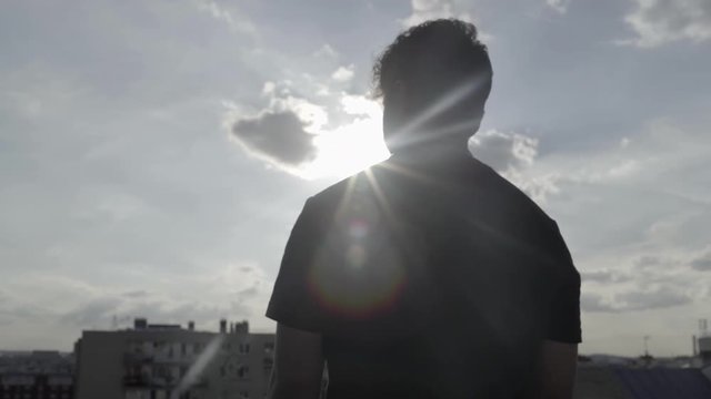 Medium shot of handsome pensive young man with curly hair and urban black tee shirt with back light and beautiful light flair and beautiful sun in cloudy sky, view of Paris rooftops in 4K