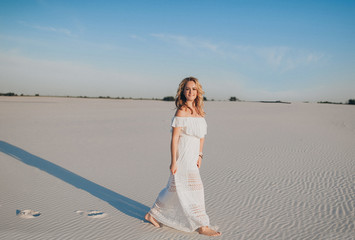 Fototapeta na wymiar A beautiful young girl blonde in a white dress walk through the sand and smiles happily. The concept of joy, ease and freedom during the vacation.