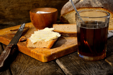 A glass of tea, bread, salt, an aluminum old kettle, a sandwich with butter, a knife on a wooden table in the kitchen.