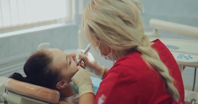 Female dentist gently drills a tooth to a patient, removes odontolith. Girl patient is sitting in the dentist's chair with an open mouth. Bright clinic with modern equipment.