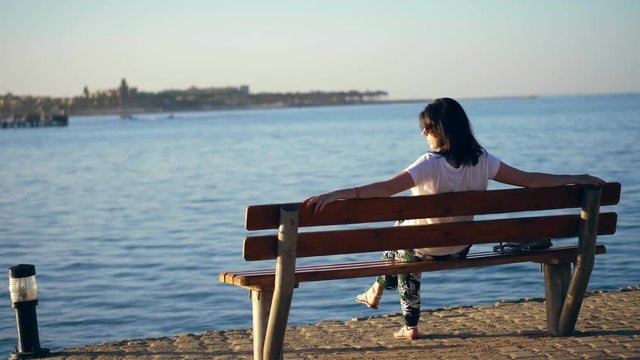 summer, sunset, embankment by the sea, a young brunette woman sit on a bench, back, admiring a beautiful sunset. lonely woman on a bench by the sea at sunset