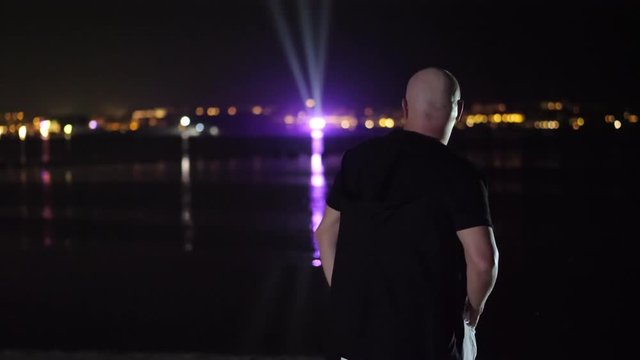 night, a bald man is dancing, view from the back. against the lights of the glowing city above the water