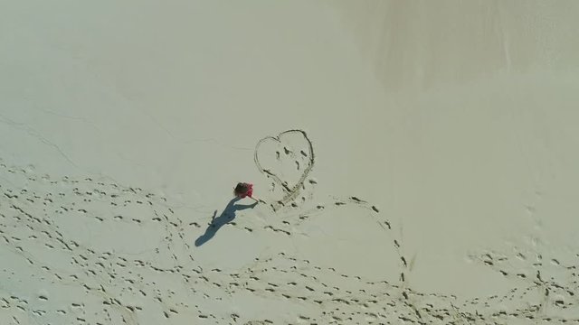 Aerial drone shot looking directly down with a woman in a red dress drawing a heart in the sand on the beach.