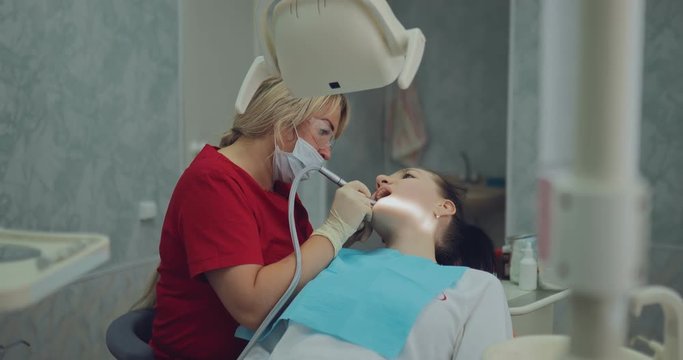 Beautiful woman dentist treats a tooth to a girl, drills a tooth. Oral hygiene procedure has finished, the drill removes the tools on the rack.