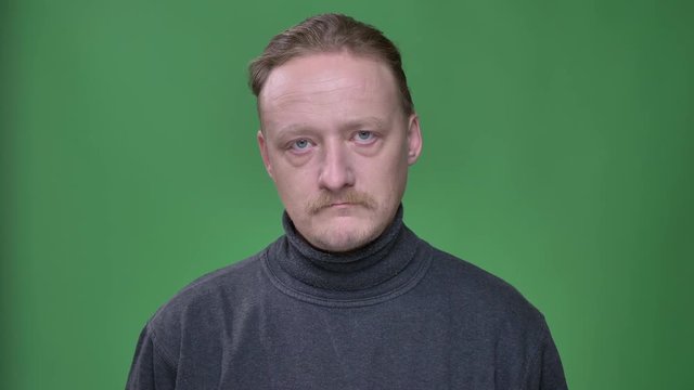 Blonde middle-aged man in gray pullover thoughtfully raising his eyes and watching into camera on green background.