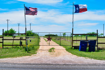 American and Texas state flags flying near countryside gate, Texas rustic star with road to the house slowly dissolving in the background.