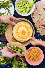Adult woman and kid, child hands holds, eats creamy chickpea Israeli Arabic hummus dip paste with tahini, oil, garlic, paprika served with fresh vegetables, salad leaves and baked pita flat breads.