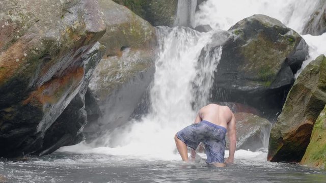 Young man in streaming water from tropical waterfall in wild jungle. Traveling man standing on rocks in flowing river stream from waterfall in rainforest.
