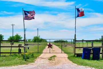American and Texas state flags flying near countryside gate, Texas rustic star with road to the house slowly dissolving in the background.