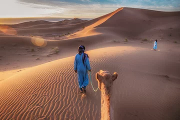 Crédence en verre imprimé Maroc Deux nomades touaregs vêtus de longues robes bleues traditionnelles, conduisent un chameau à travers les dunes du désert du Sahara au lever du soleil au Maroc.