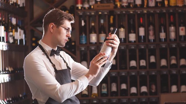 Handsome and young smiling brown haired sommelier with a bottle of wine on the background of dark wine house with shelves of bottles with alcohol
