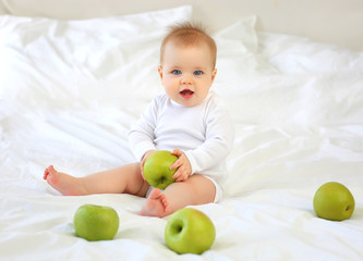 Charming blue-eyed 9-month-old kid with funny hair sitting on the bed with green apples. White clothes, blond hair.