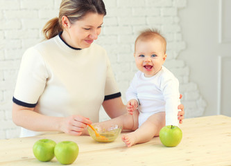 Fototapeta premium 9 month old charming baby sits on the table and mom feeds him with a spoon of applesauce. Blonde hair, blue eyes.
