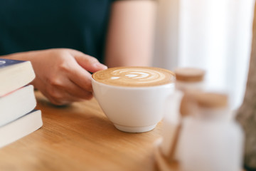 Fototapeta na wymiar Closeup image of a woman's hand holding a white cup of hot coffee with books on wooden table