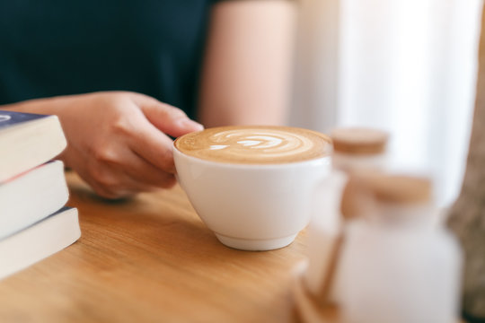 Closeup image of a woman's hand holding a white cup of hot coffee with books on wooden table