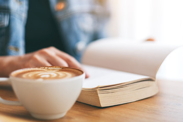 Closeup image of a woman holding and reading a vintage novel book with coffee cup on wooden table