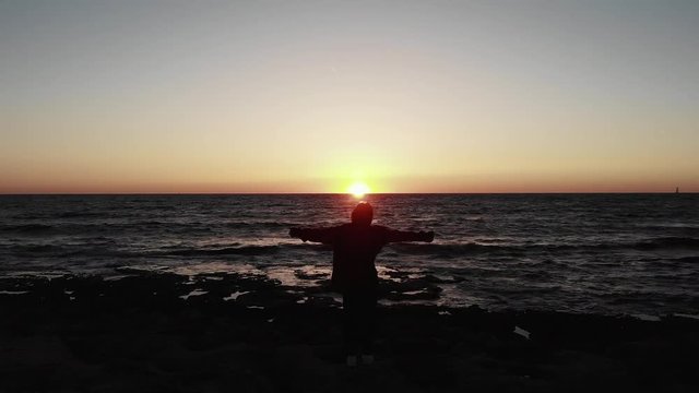 silhouette of young female woman standing on the rocky beach with hands widespread to sides on sunset over the wavy sea with sun road. Aerial drone view