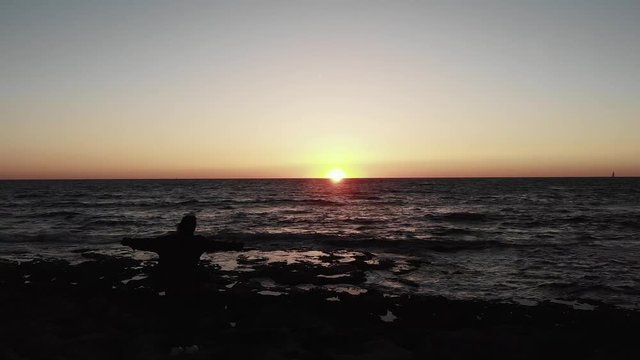 Black silhouette of woman on rocky sea beach hands spread wite during sunset over ocean with waves hitting beach. Aerial drone shot