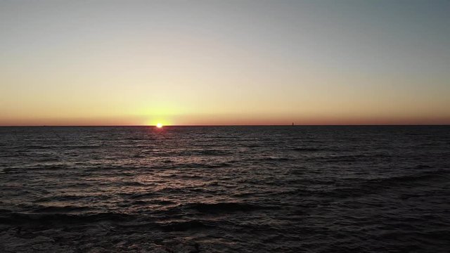 Sunset over ocean with waves hitting beach and small yacht on horizon. Sunset with sun road on wavy sea ocean.