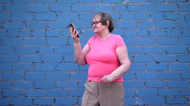 elderly woman in pink T-shirt and glasses dances with new smartphone model and enjoys beautiful quality sound in headphones