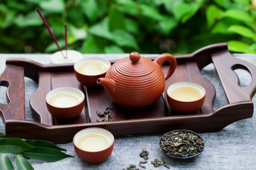 Green tea in tea pot and bowls, cups on a wooden tray. Outdoor background.