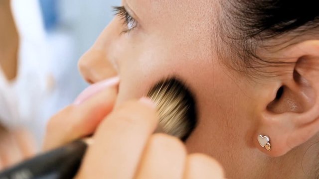Slow motion video of young beautiful fashion model sitting next to big mirror with bulbs while professional makeup artist is applying makeup on her face
