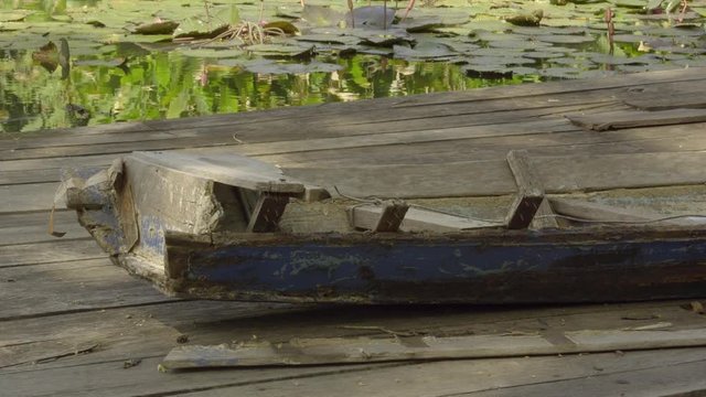 Remains of painted, wooden long boat, broken into pieces on wooden plank walkway alongside pond with floating lily pads on the surface in sunlight.