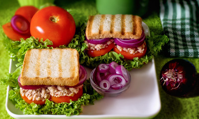 Tuna sandwiches on a square china dish, tomatoes, lettuce, red onion bulb. Two fish sandwiches. White-green background, cloth napkins. Selective focus.