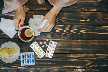ill girl with a flu is sitting at the wooden table with pills and tea. Runny nose and cold. Weak...