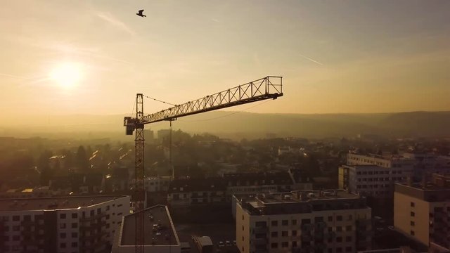 Aerial drone view of construction site crane over city at sunset with mountains in the background.