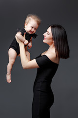 Stylish young mother in black dress holds baby daughter in studio on gray background