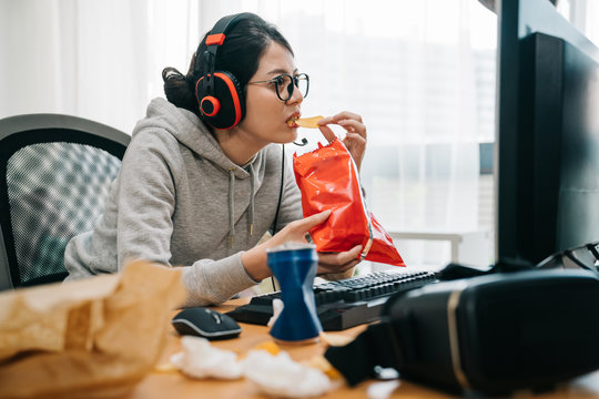 emotional funny college girl enjoying fast food during using computer in headphones. woman having summer break playing online games. young female homebody glasses staring at monitor screen eat chips