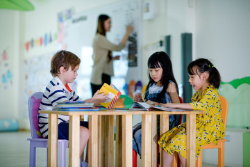 group of international kids in preschool enjoy reading books with teacher watching in background