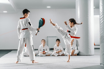 Caucasain boy and girl in doboks having taekwondo training at gym. Girl kicking while boy holding kick target. In background their friend sitting with legs crossed and watching them.