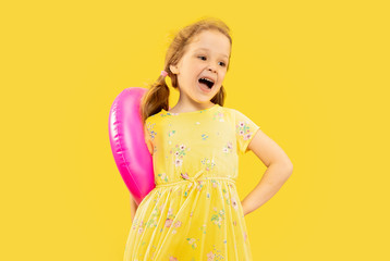 Beautiful emotional little girl isolated on yellow background. Half-lenght portrait of happy child wearing a dress and holding rubber pink flamingo. Concept of summer, human emotions, childhood.