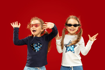 Beautiful emotional little girl isolated on red background. Half-lenght portrait of happy sisters or friends in red and black sunglasses. Concept of facial expression, human emotions, childhood.